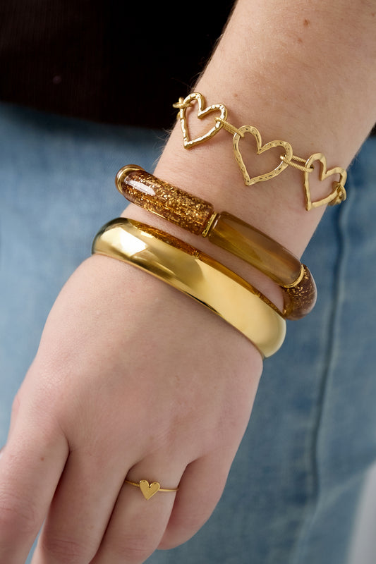 Woman's wrist adorned with three bracelets including a glitter resin bracelet, gold heart link bracelet, and gold cuff, plus a dainty heart ring