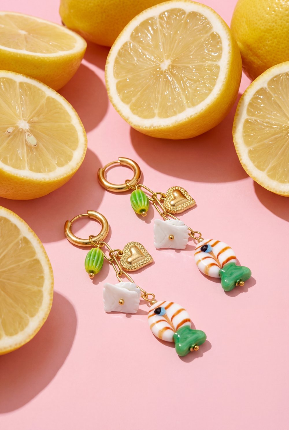 Gold-tone dangle earrings featuring charms of hearts, white squares, and fish, set against a pink background with lemons.
