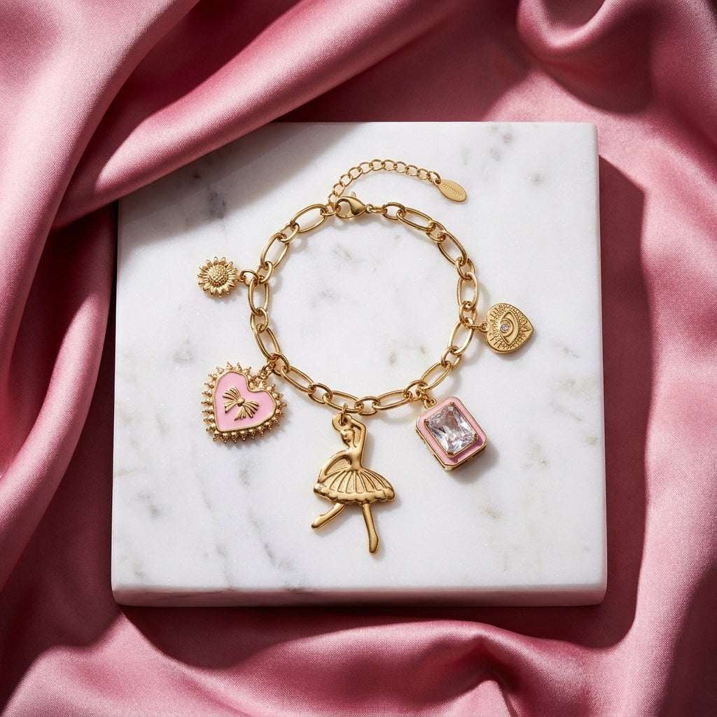 Gold chain charm bracelet with pink enamel heart, ballerina, crystal, and evil eye charms on a white marble base.