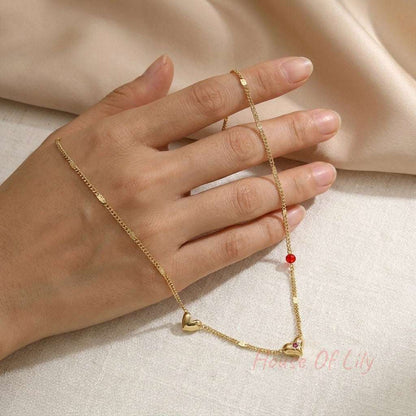 Gold chain necklace on a hand featuring small gold hearts, a red bead, and a tiny embedded pink stone.
