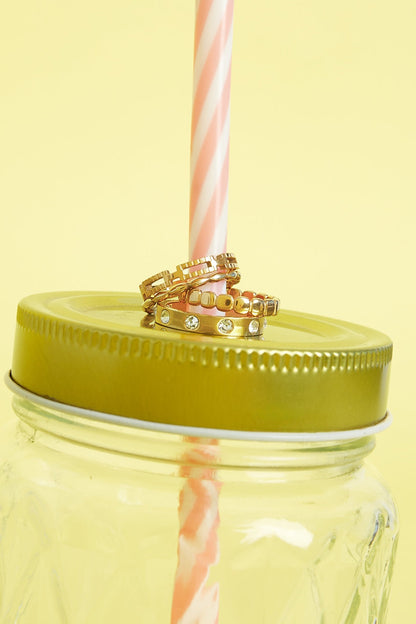 Three gold rings stacked on a golden lid of a jar with a pink and white striped straw against a yellow background