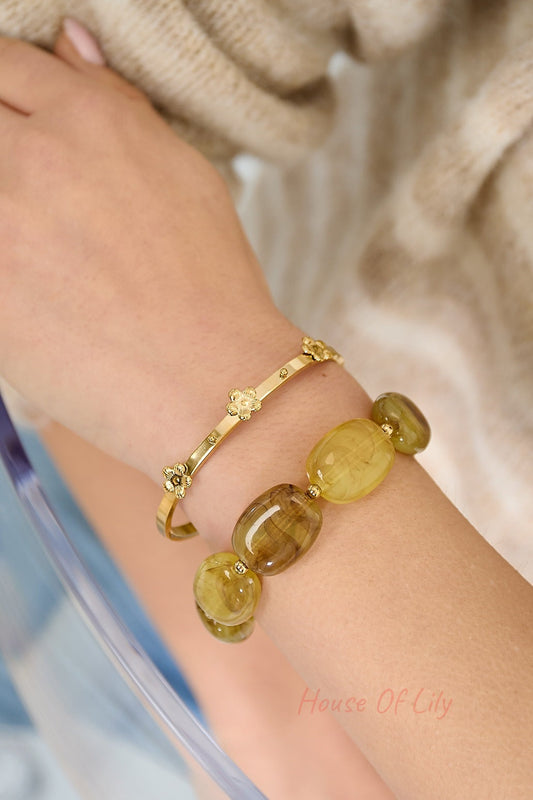 Wrist adorned with the gold floral detail bangle paired with a chunky yellow-green beaded bracelet, with cozy beige sweater background