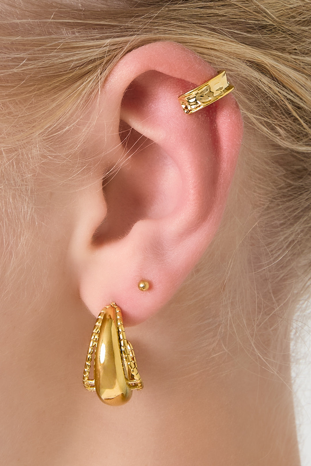 Close-up of a woman's ear wearing a hammered gold ear cuff on the cartilage and layered gold earrings on the lobe