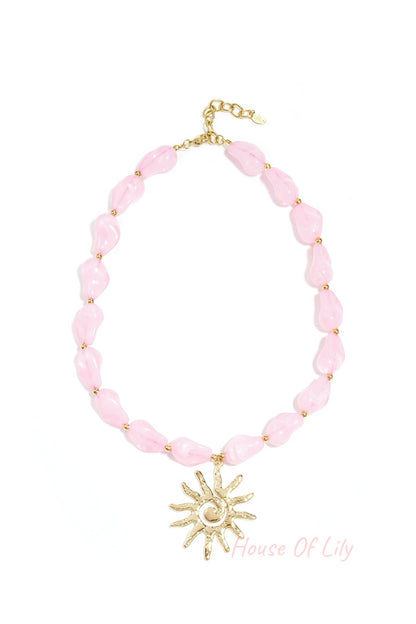 Soft pink acrylic beaded necklace with gold sun pendant displayed on a white background
