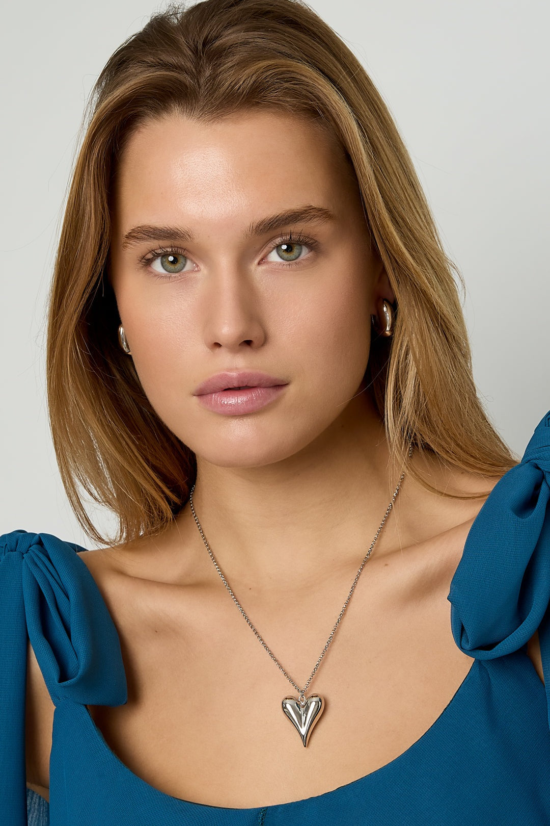 Close-up portrait of a woman wearing a silver-tone puffy heart pendant necklace with a blue off-shoulder top