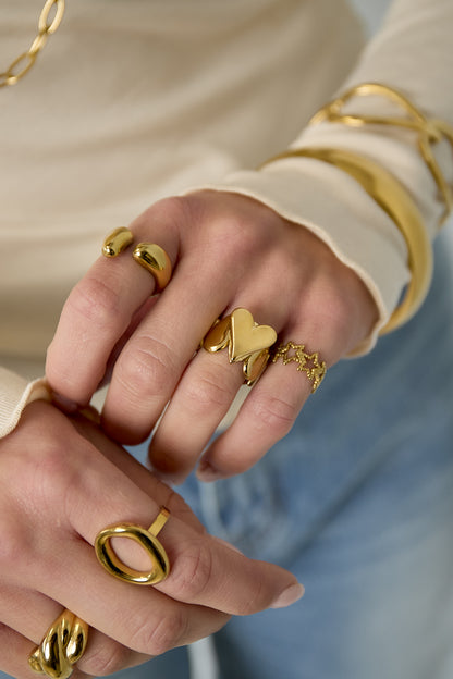 Close-up of female hand wearing gold heart statement ring with other gold rings and bracelets.