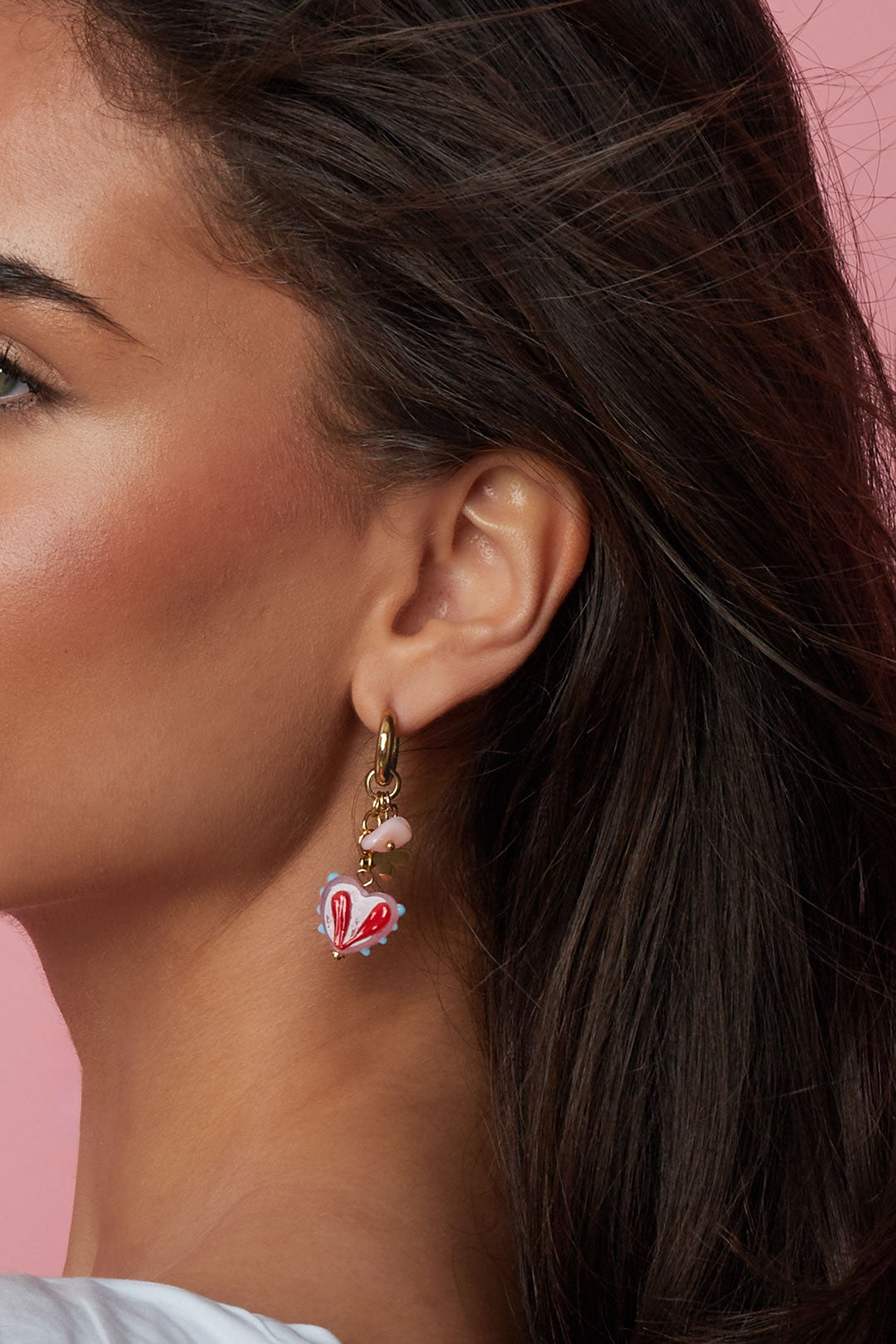 Closeup side profile of model wearing gold hoop earrings with pink heart charm on pink backdrop
