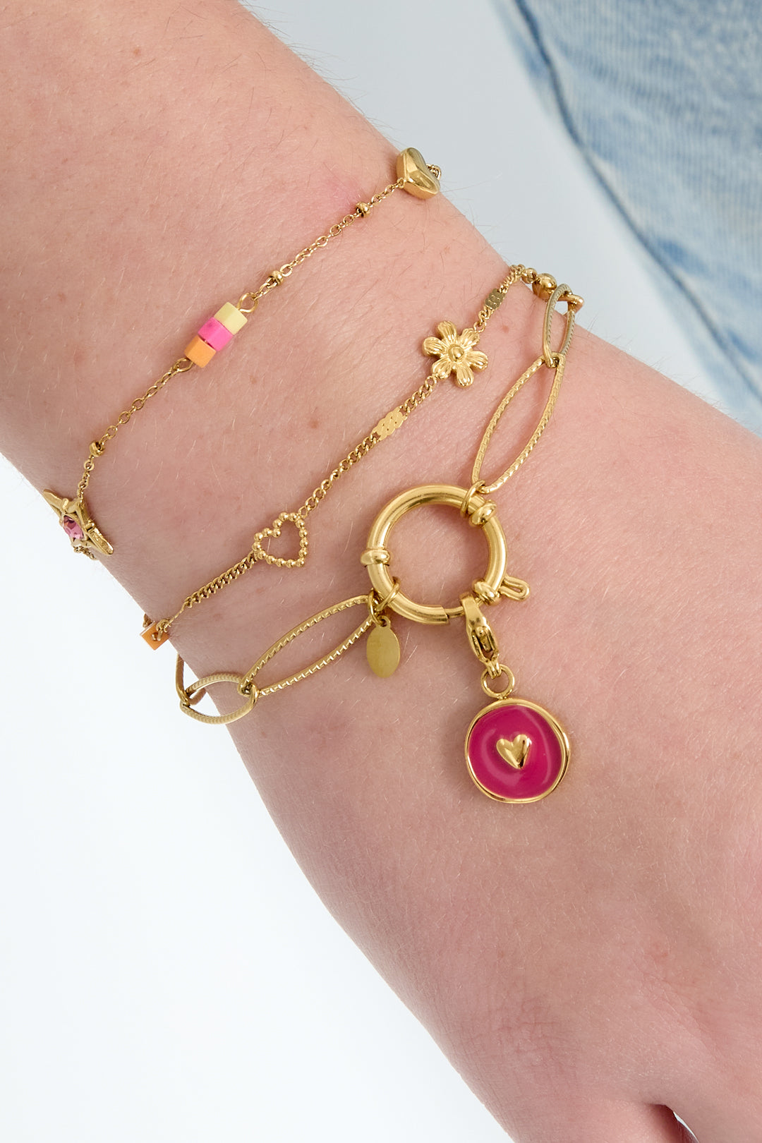 Close-up of a wrist adorned with layered gold bracelets, one featuring a gold link chain and toggle clasp with a pink charm, styled with denim