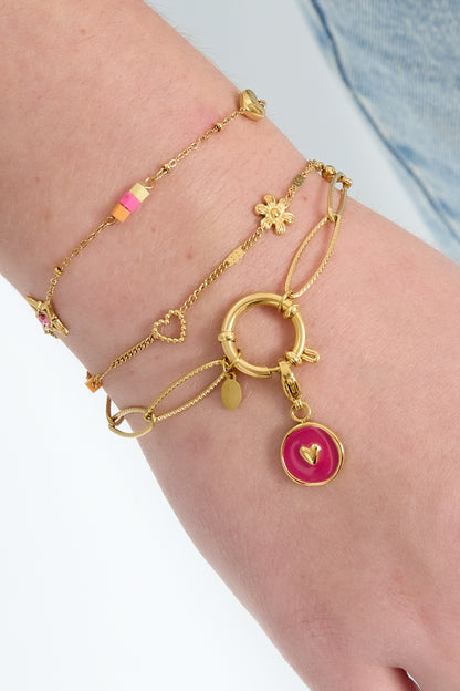Close-up of a wrist adorned with layered gold bracelets, one featuring a gold link chain and toggle clasp with a pink charm, styled with denim
