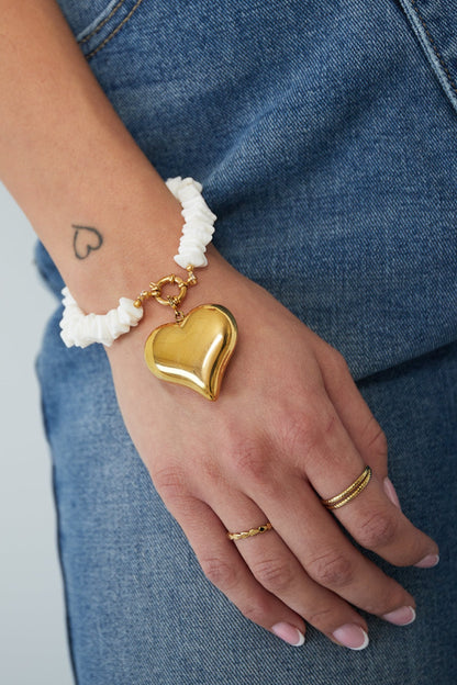 Close-up of hand wearing white shell beaded bracelet with large gold heart charm and gold toggle clasp against denim jeans