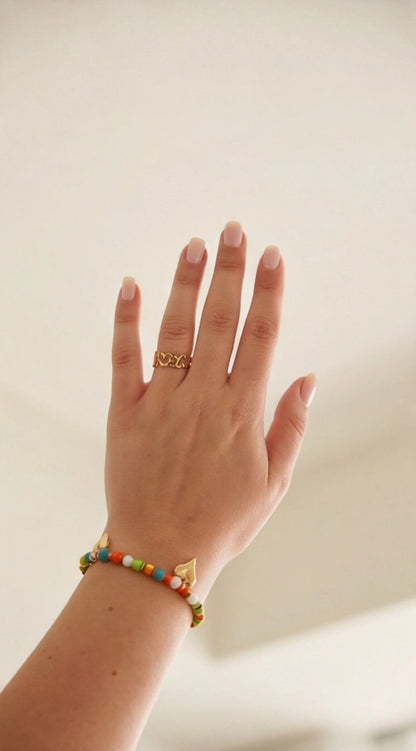 Hand wearing a gold ring and colorful beaded bracelet on a plain background