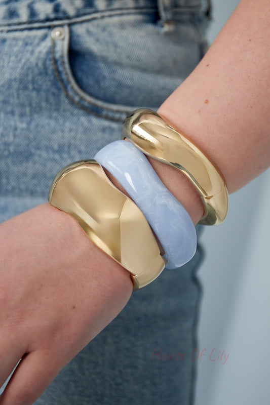 Close-up of a wrist wearing two golden sculpted bangles and one light blue resin bangle, styled with denim jeans