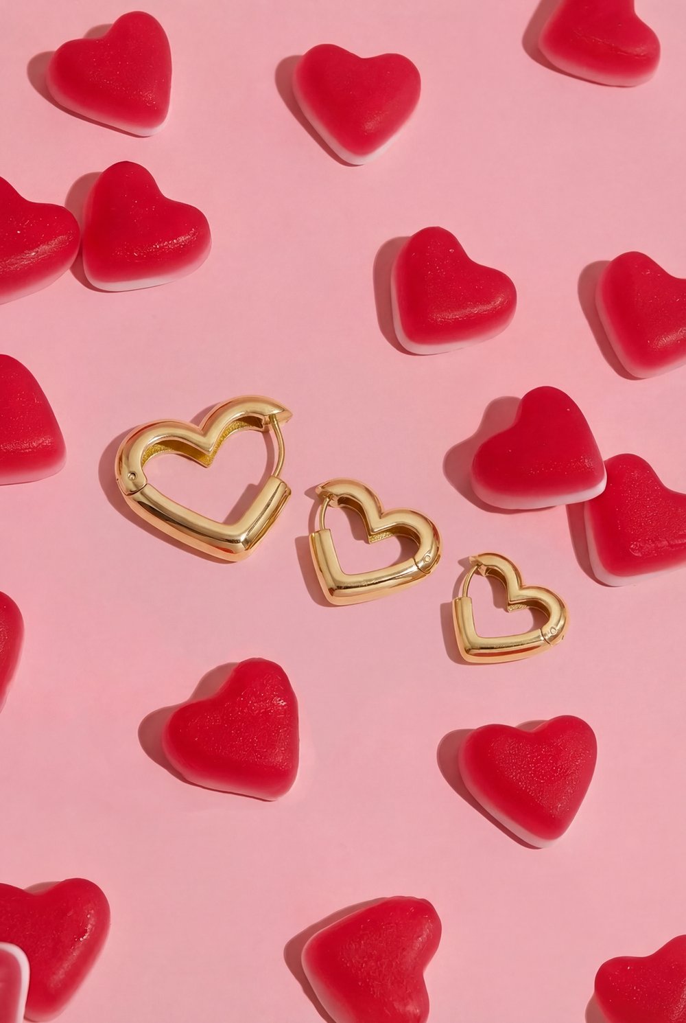 Three gold heart-shaped hoop earrings of varying sizes, scattered on a pink surface with red gummy heart candies.