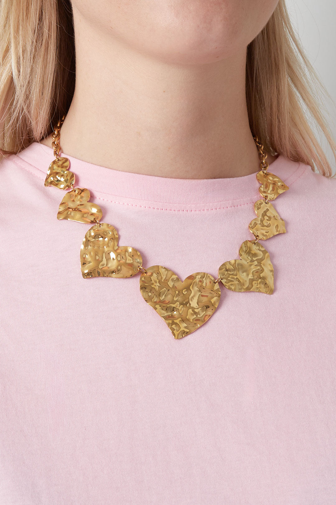 Close-up of woman wearing chunky hammered gold heart necklace over a pink shirt, showing fit and texture