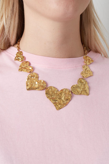 Close-up of woman wearing chunky hammered gold heart necklace over a pink shirt, showing fit and texture
