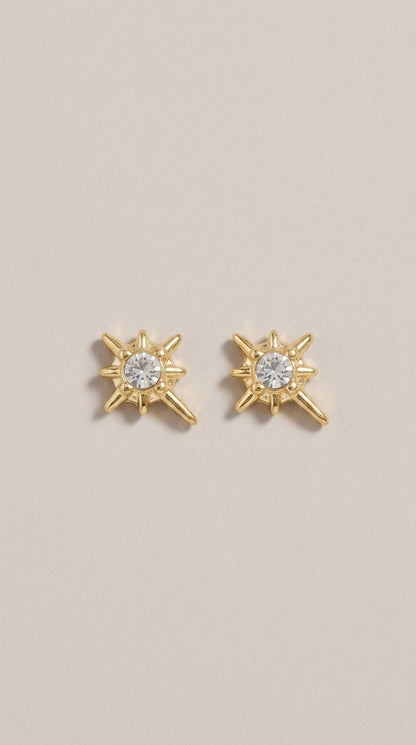 Gold stud earrings featuring a central crystal surrounded by a starburst design with one elongated ray.
