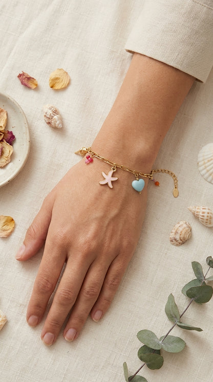 Coastal Charm Stack gold chain bracelet featuring a light pink starfish, blue heart charm, and red beads on linen.