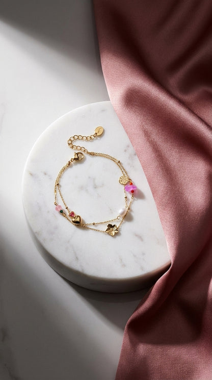 Gold charm bracelet stack with a heart, flower, pearl, and pink stones on a marble surface with pink silk.