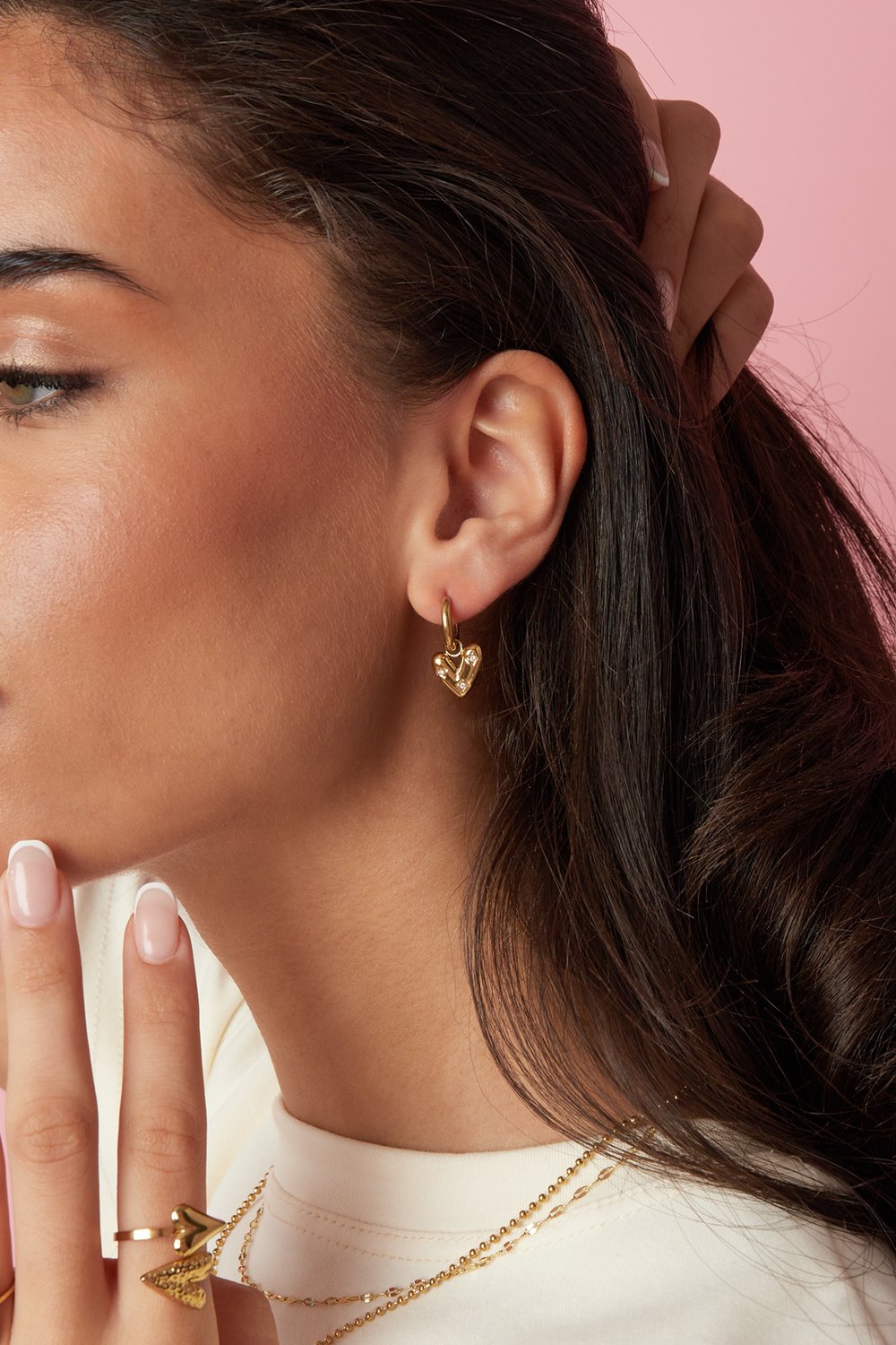 Gold Main Character Heart hoop earrings featuring small, sparkling inlaid crystals, worn on a model against pink.