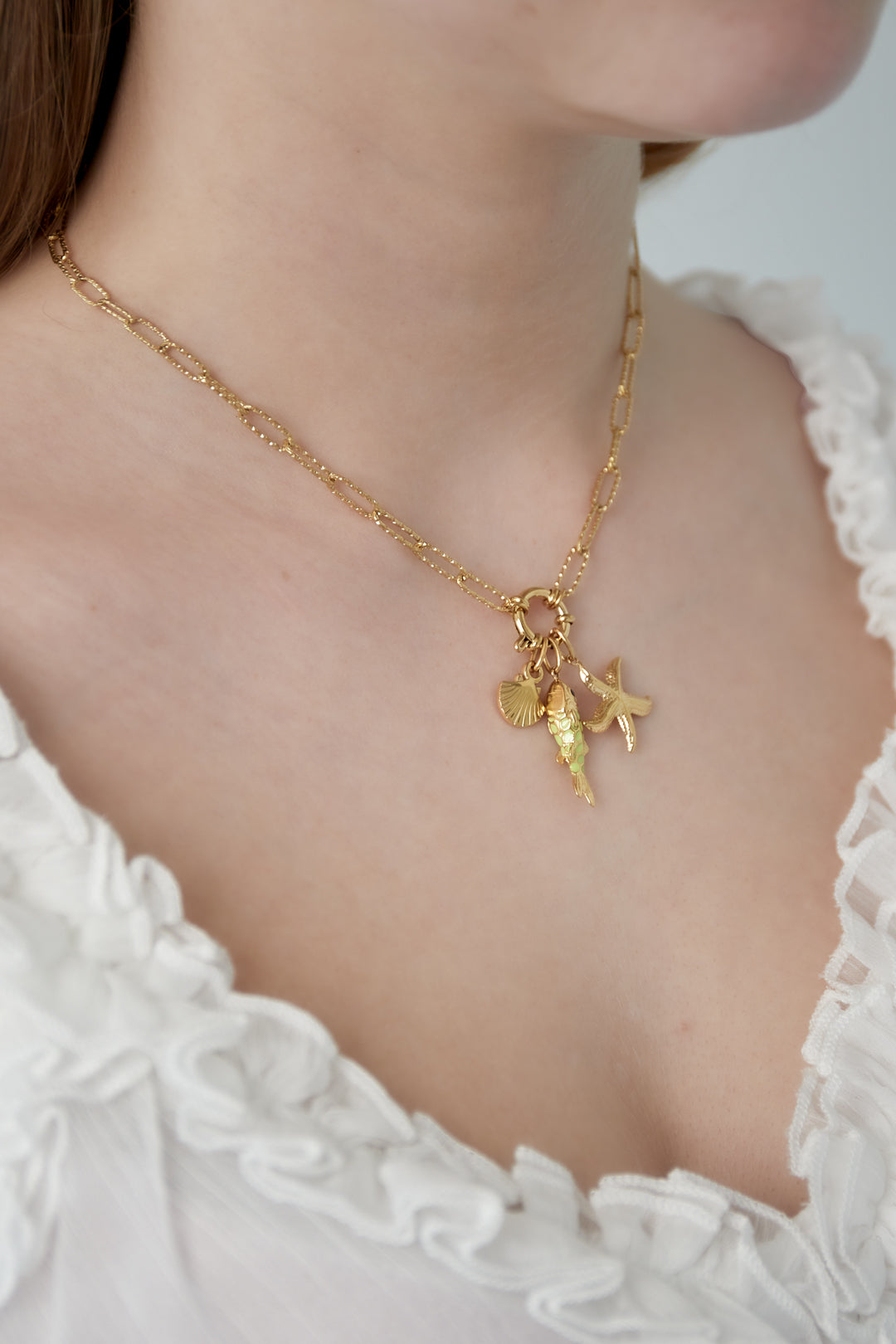 Close-up of the golden ocean charm necklace worn by a woman dressed in a white frilly top, showcasing the starfish, fish and shell charms