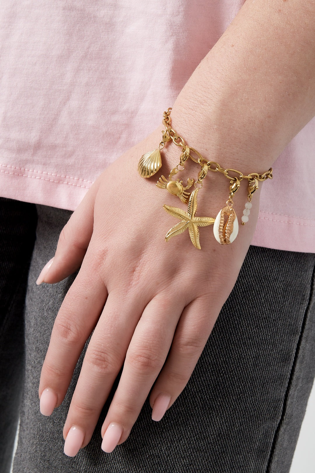 Close-up of female wrist wearing the gold charm bracelet with ocean-themed charms, styled with casual pink top and dark jeans