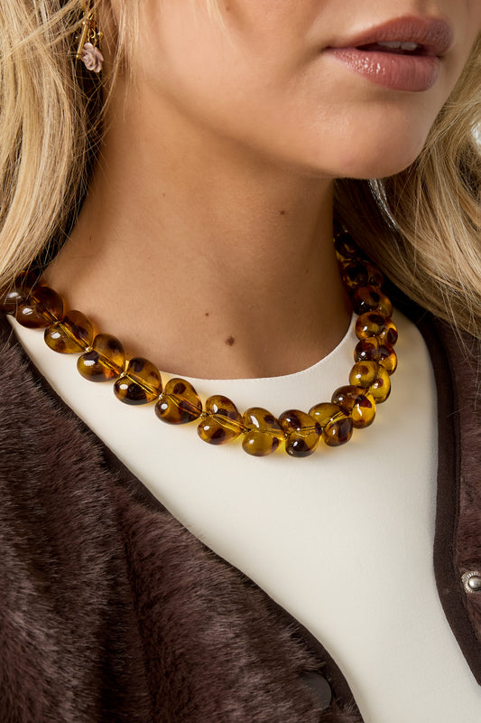 Detailed view of amber heart bead necklace shown on woman wearing cream top and brown vest
