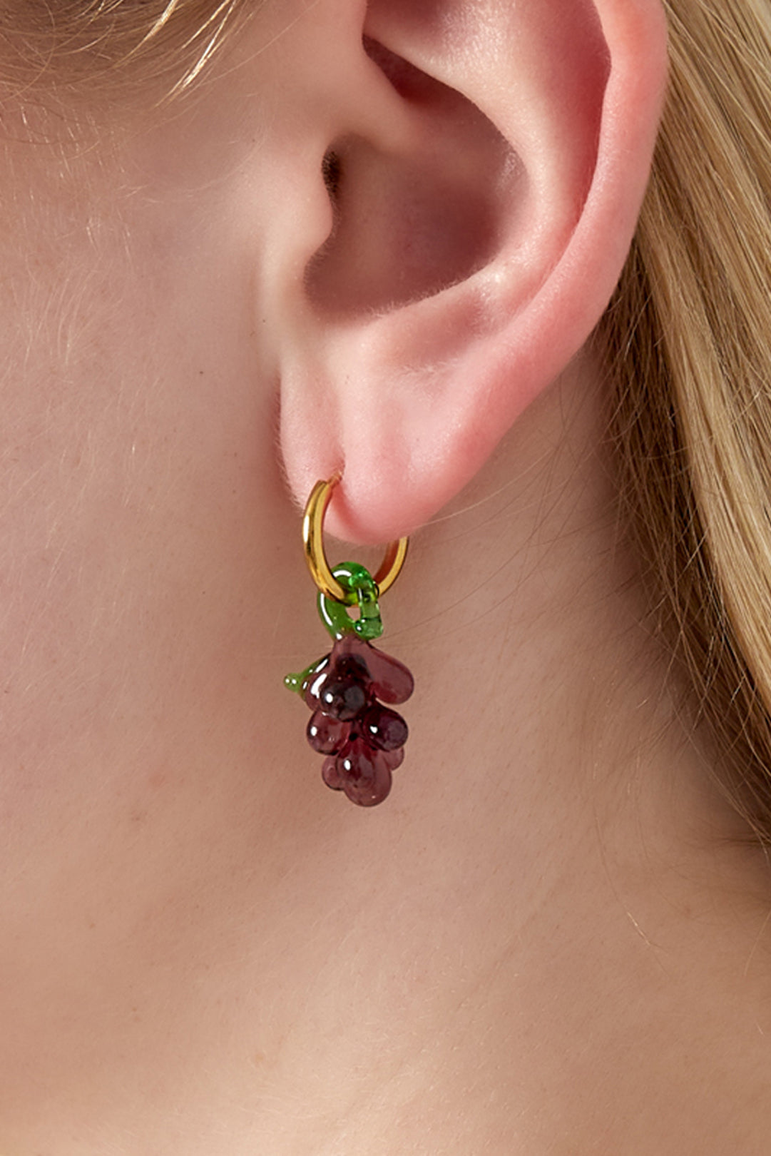 Close-up of a gold hoop earring with a purple glass grape charm hanging, worn on an ear
