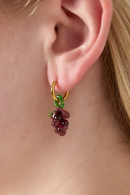 Close-up of a gold hoop earring with a purple glass grape charm hanging, worn on an ear