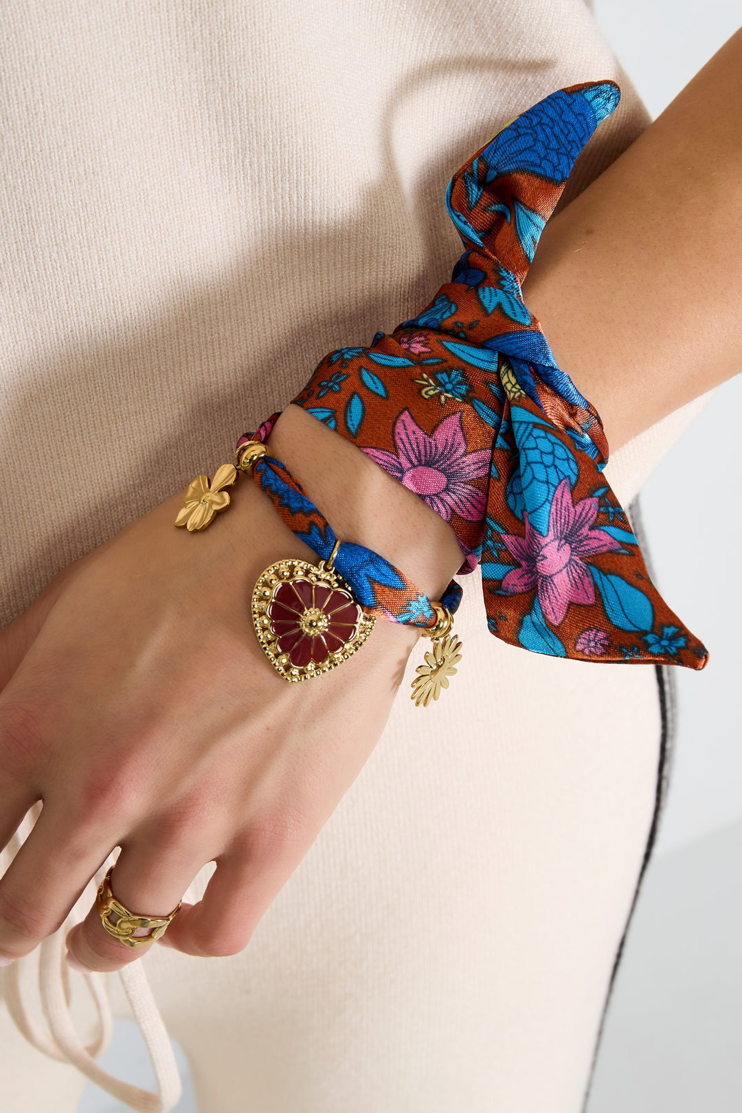 Model wearing a floral charm bow bracelet on wrist, paired with cream knit outfit and gold rings, highlighting soft fabric and gold floral charms