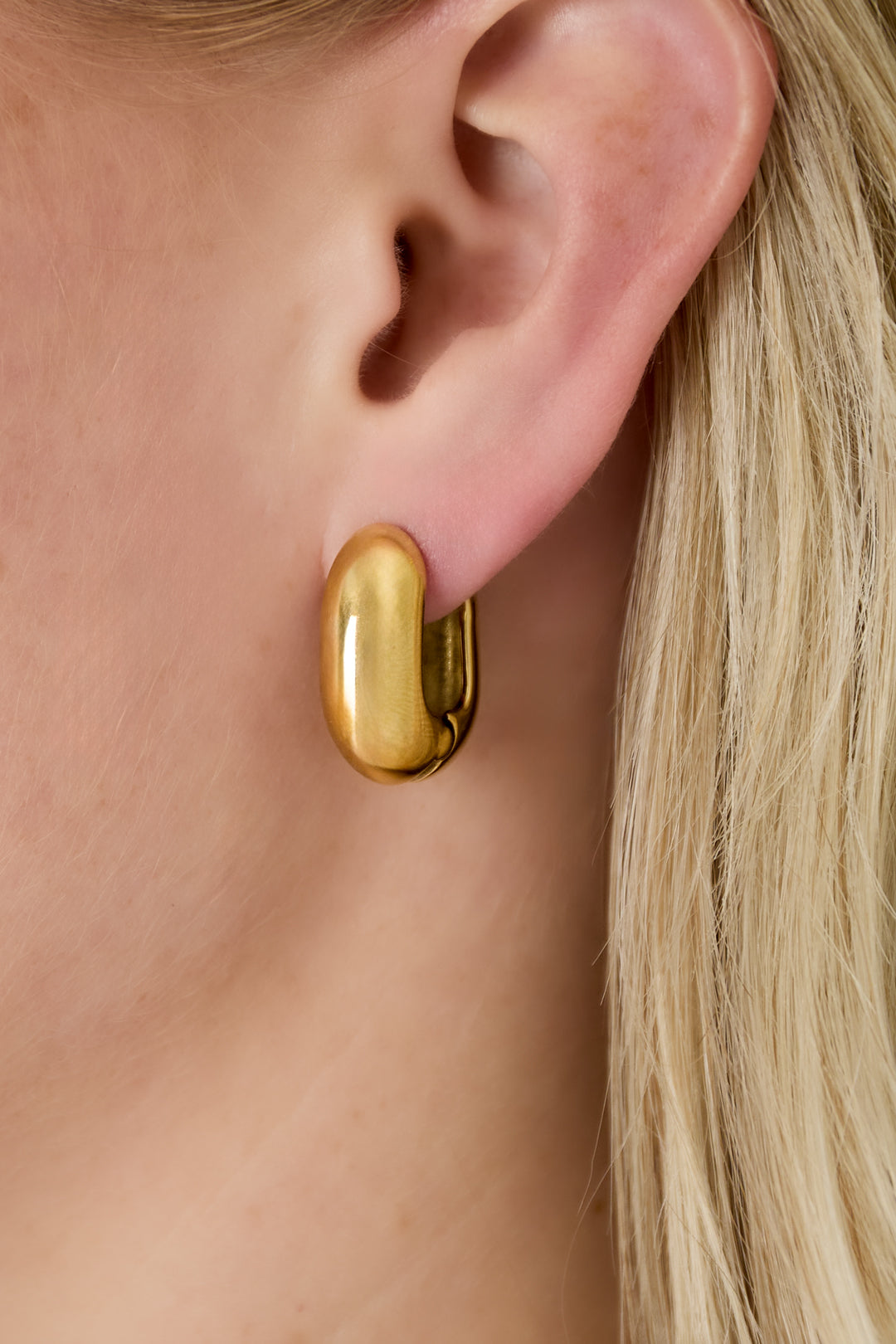 Close-up of gold chunky oval hoop earring worn on a woman's ear highlighting smooth polished design