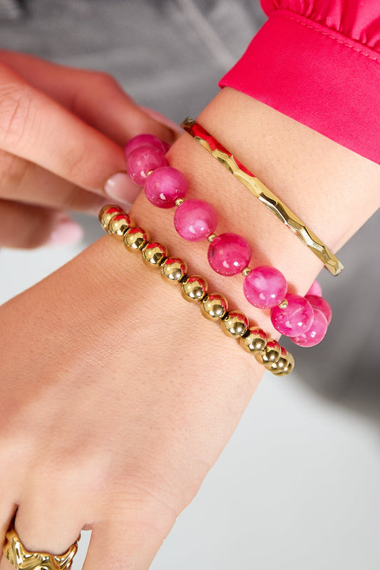 Close-up of wrist wearing three bracelets: pink bead bracelet, gold bead bracelet, and gold hammered bangle, paired with pink sleeve