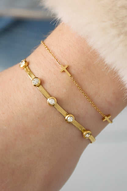 Close-up photo showing two gold bracelets on wrist: one with clear rhinestones and one with small gold crosses, styled with fur cuff