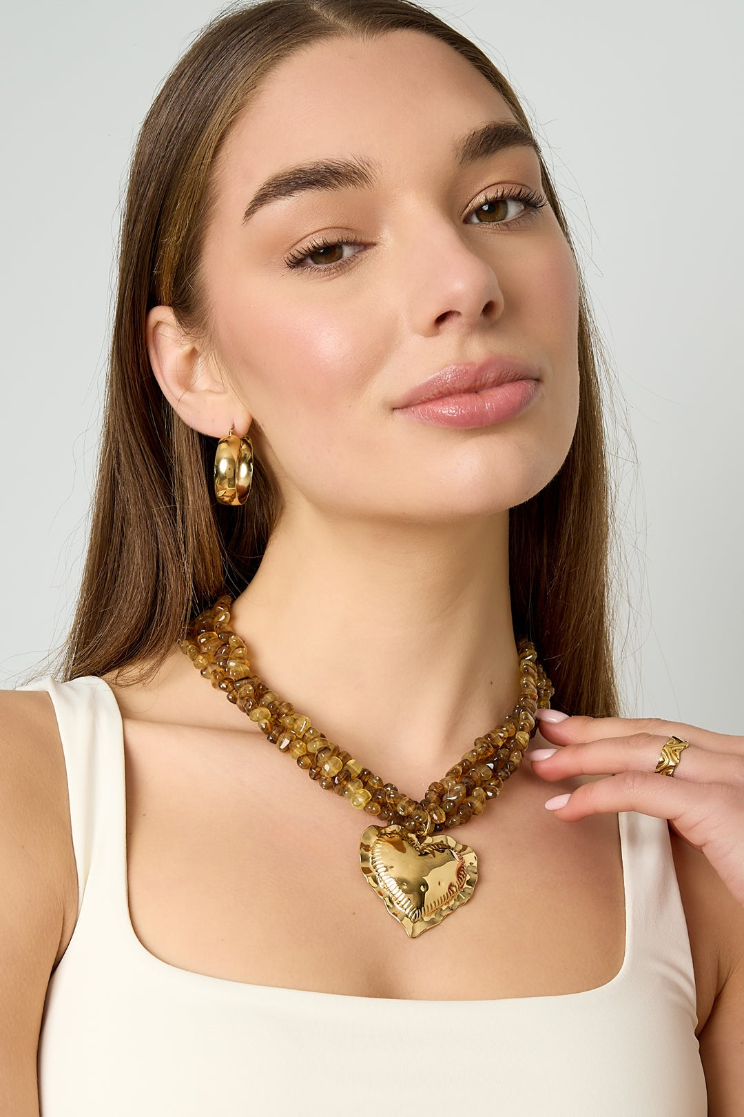 Model wearing the twisted amber bead necklace with gold heart pendant along with gold hoop earrings and white tank top