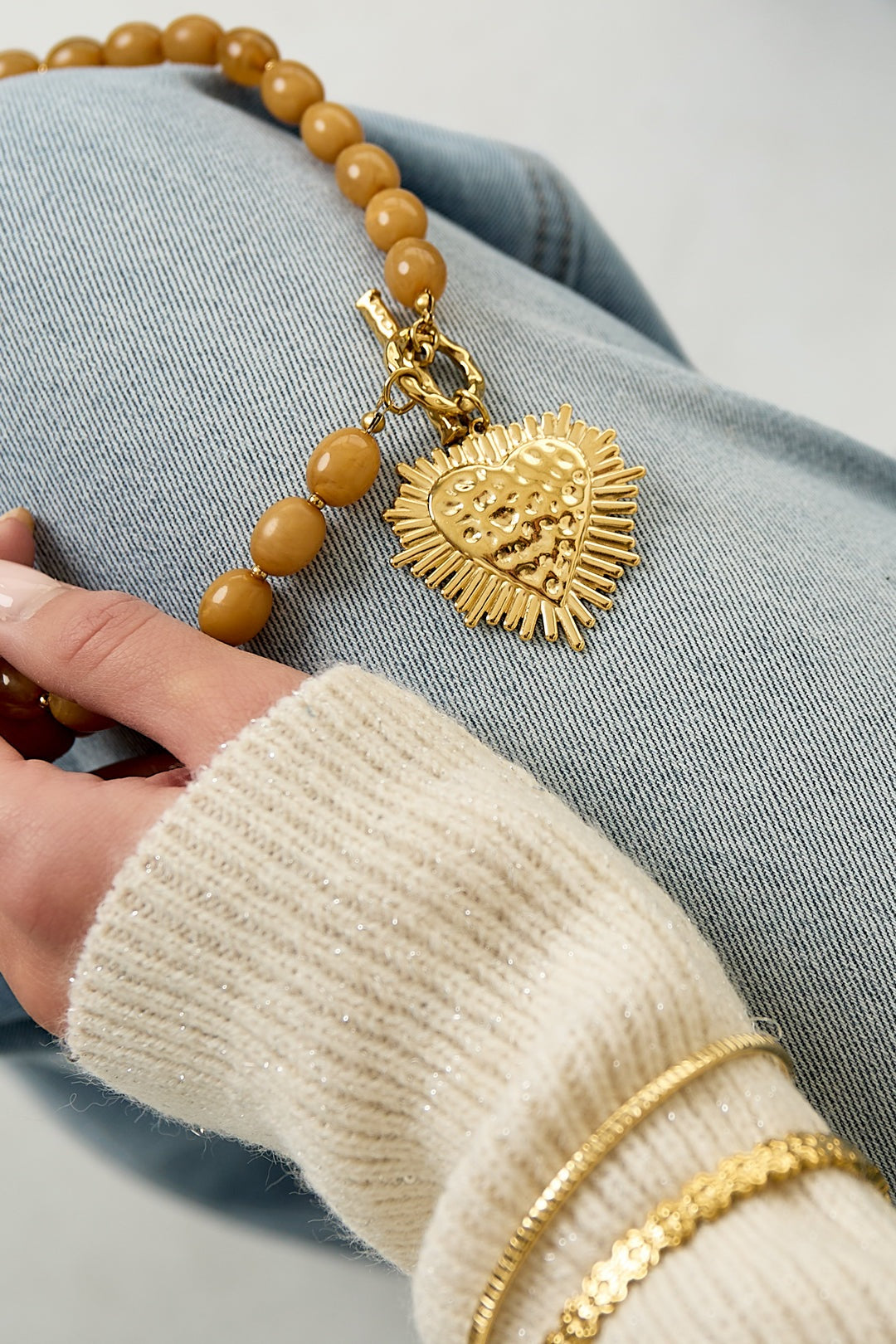 detail of nude beige beaded necklace heart pendant held by hand with gold bracelets on wrist and denim background