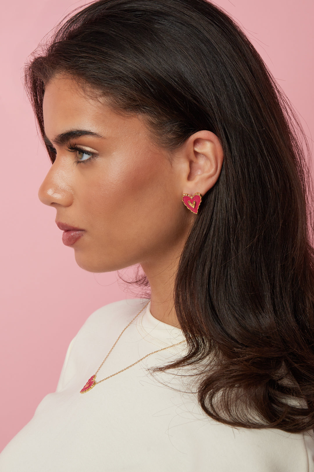 Model wears Puffy Heart Vibes gold-tone earrings with pink enamel heart centers against a pink background.