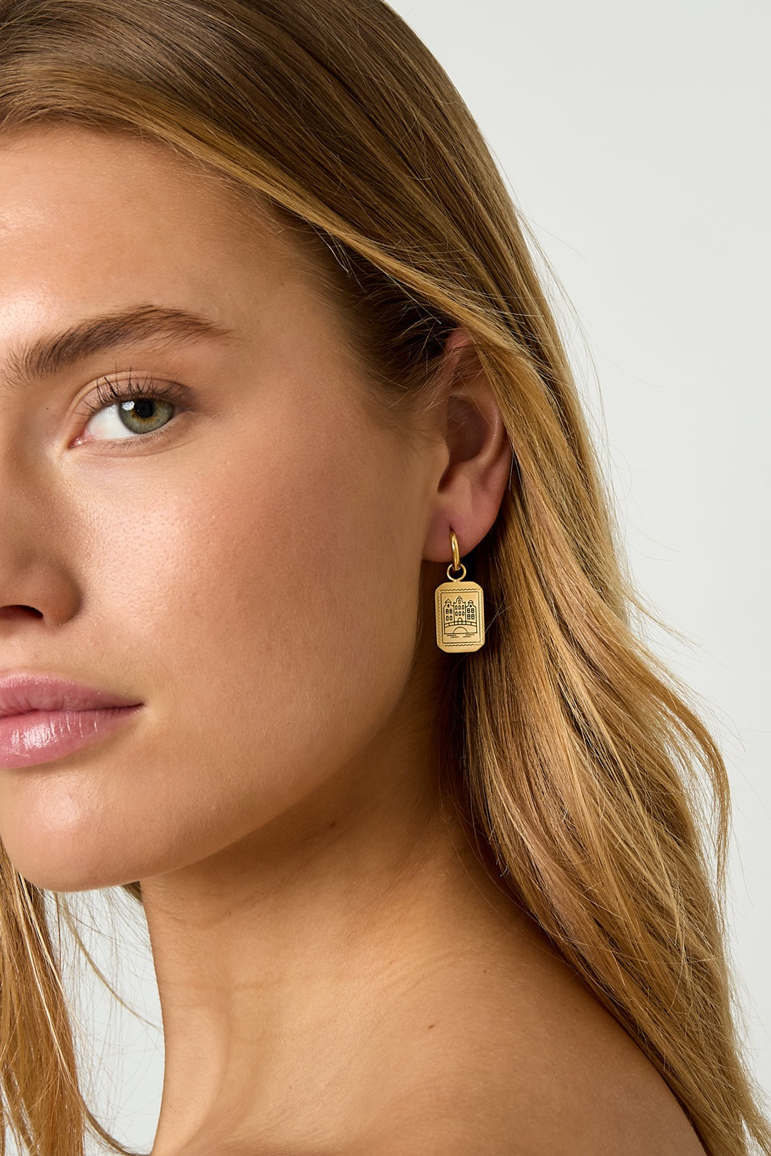 Side profile close-up of a woman wearing one gold rectangular pendant earring etched with an Amsterdam canal house.