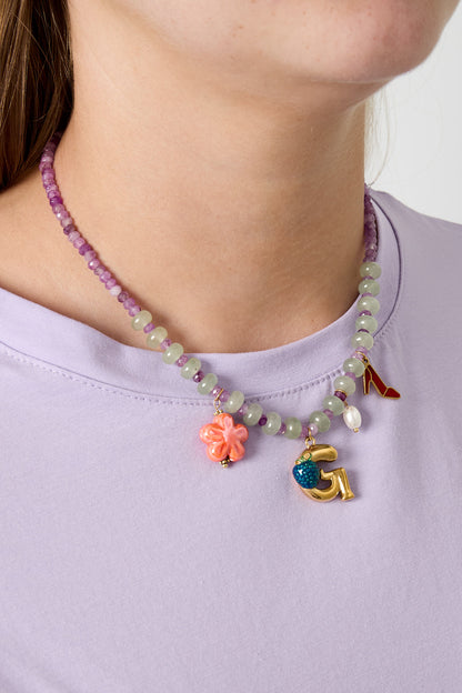 Model wearing a mixed stone beaded necklace with purple and translucent green beads, decorated with a coral flower, gold letter G with blue berry charm, pearl, and red shoe charms