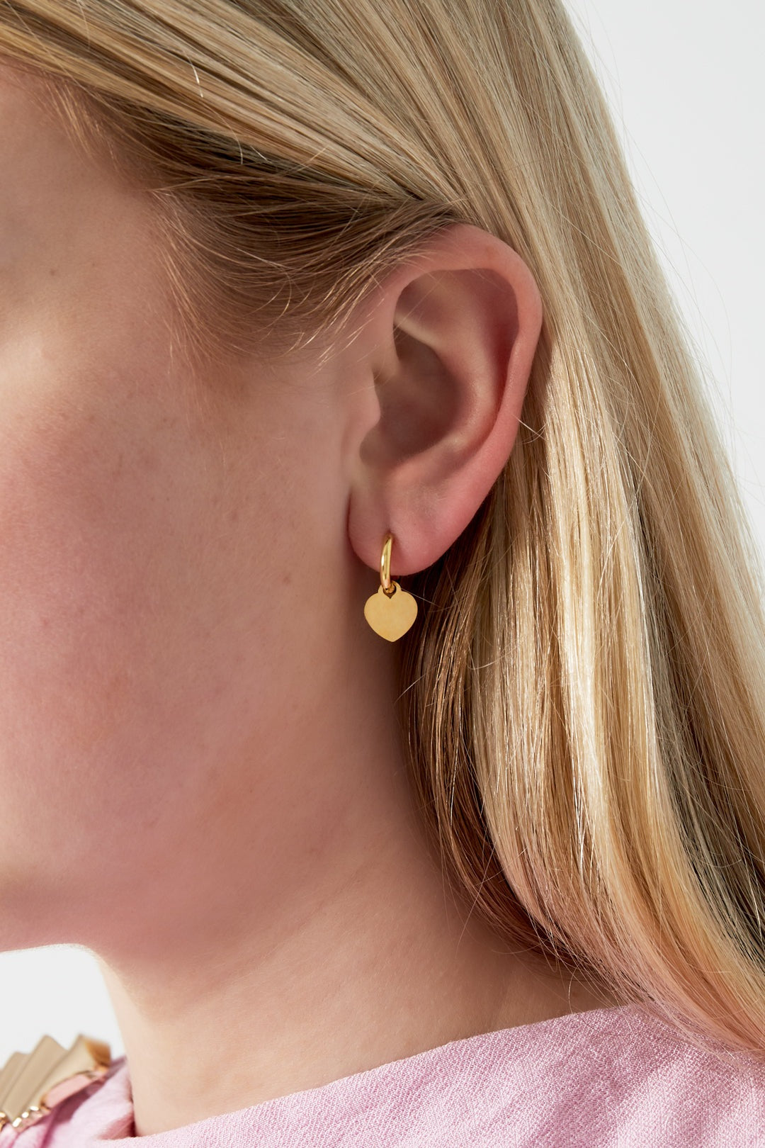 Close-up side profile of woman wearing gold heart drop hoop earrings with blonde hair and pink top