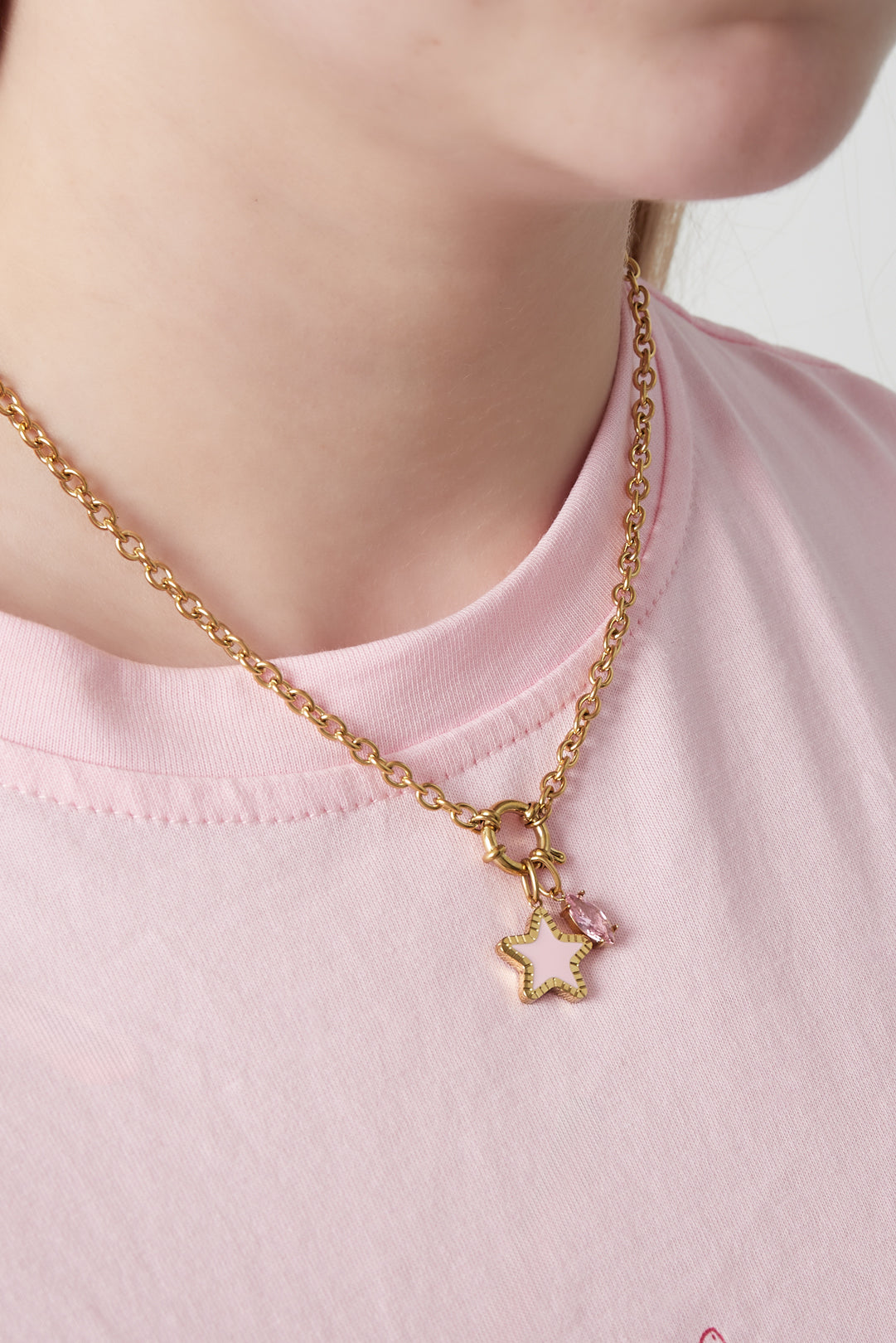Close-up of the gold-plated charm necklace worn over a soft pink t-shirt, highlighting the pink star and pink gemstone charms hanging from a toggle clasp