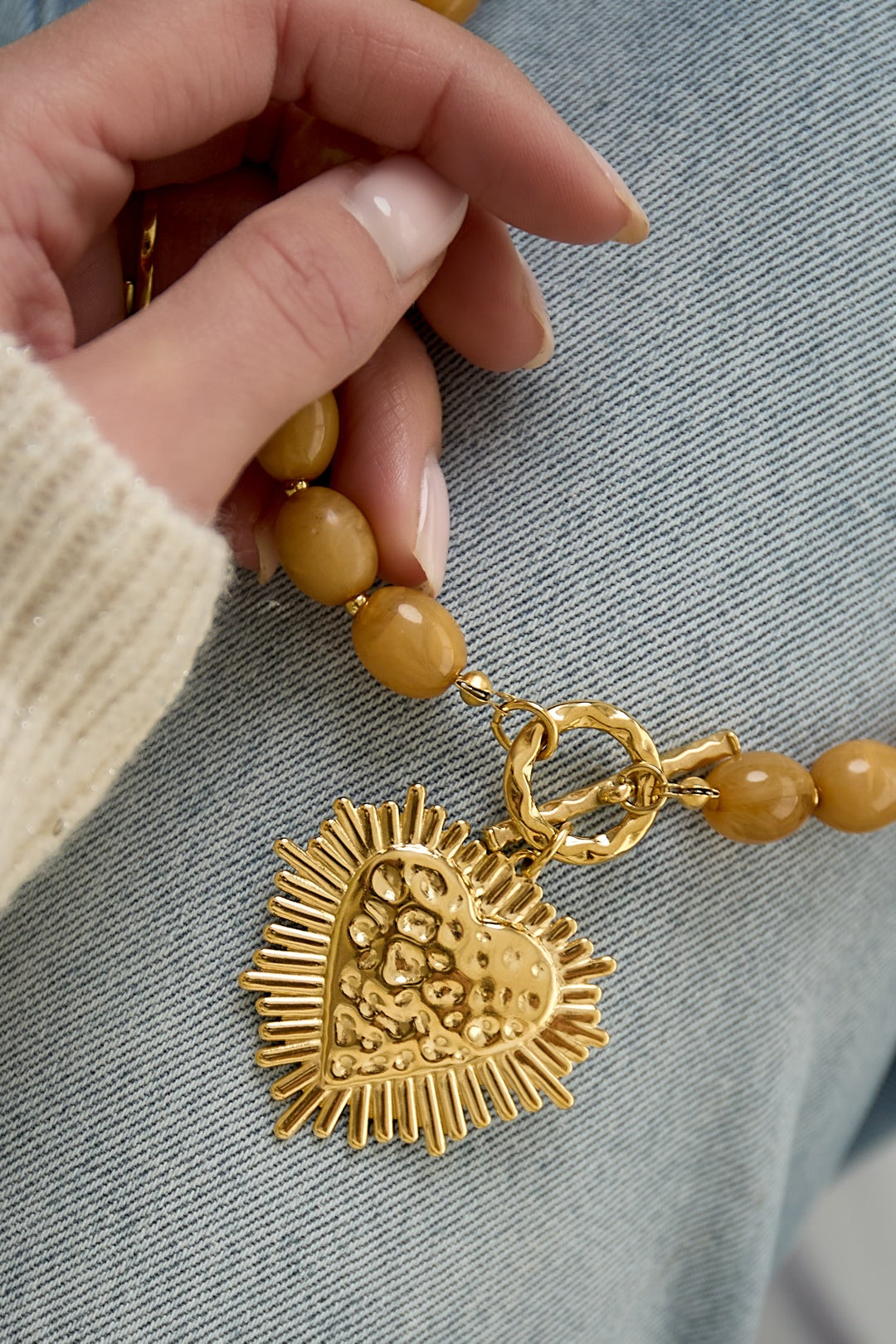 close-up of hand holding nude beige beaded necklace showing gold textured heart pendant and toggle clasp on denim