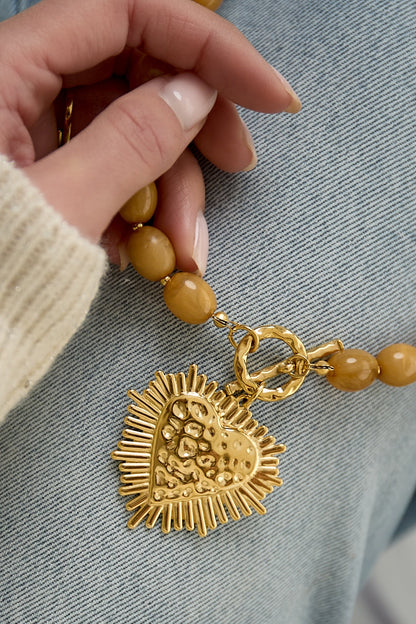 close-up of hand holding nude beige beaded necklace showing gold textured heart pendant and toggle clasp on denim