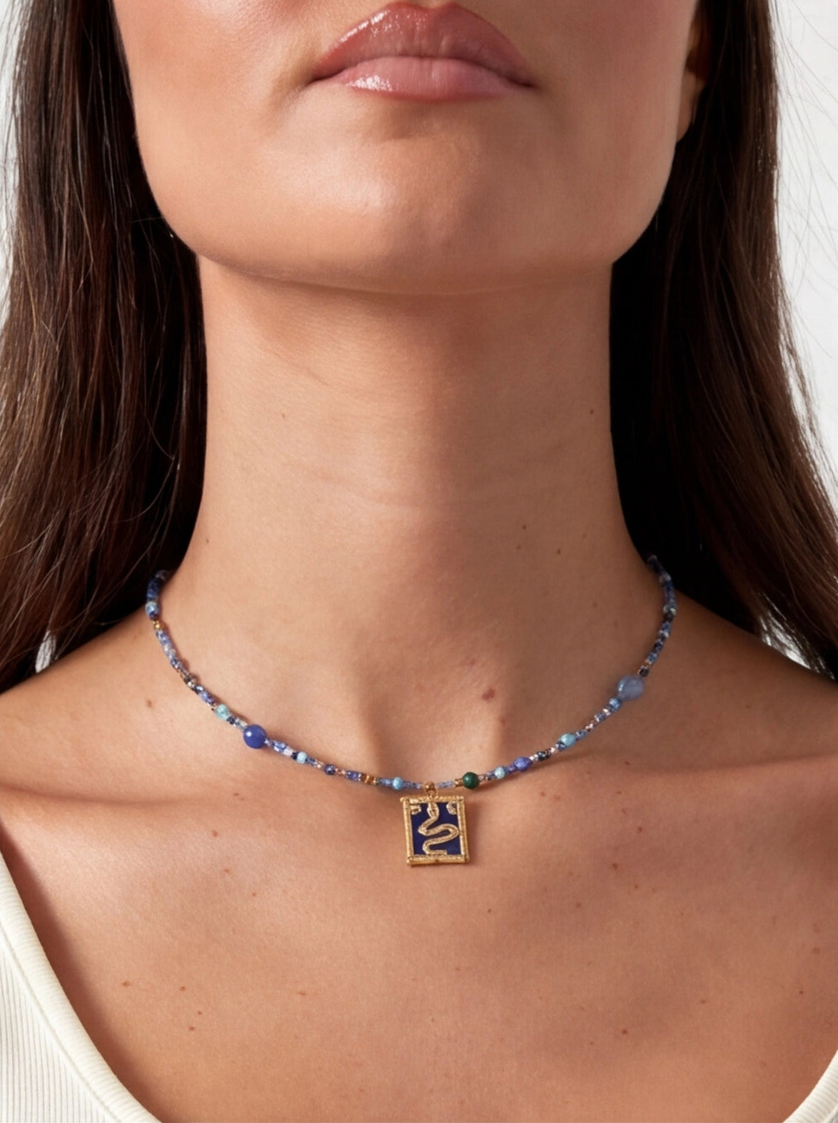 Blue beaded necklace with a gold rectangular pendant featuring a snake design, worn around the neck.