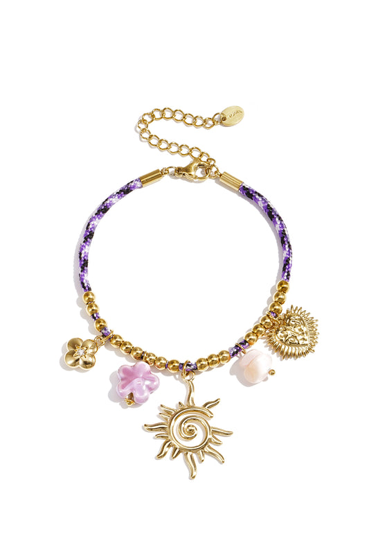 Close-up of a purple braided cord charm bracelet with gold beads and assorted charms including a sunburst, flowers, and heart pendant