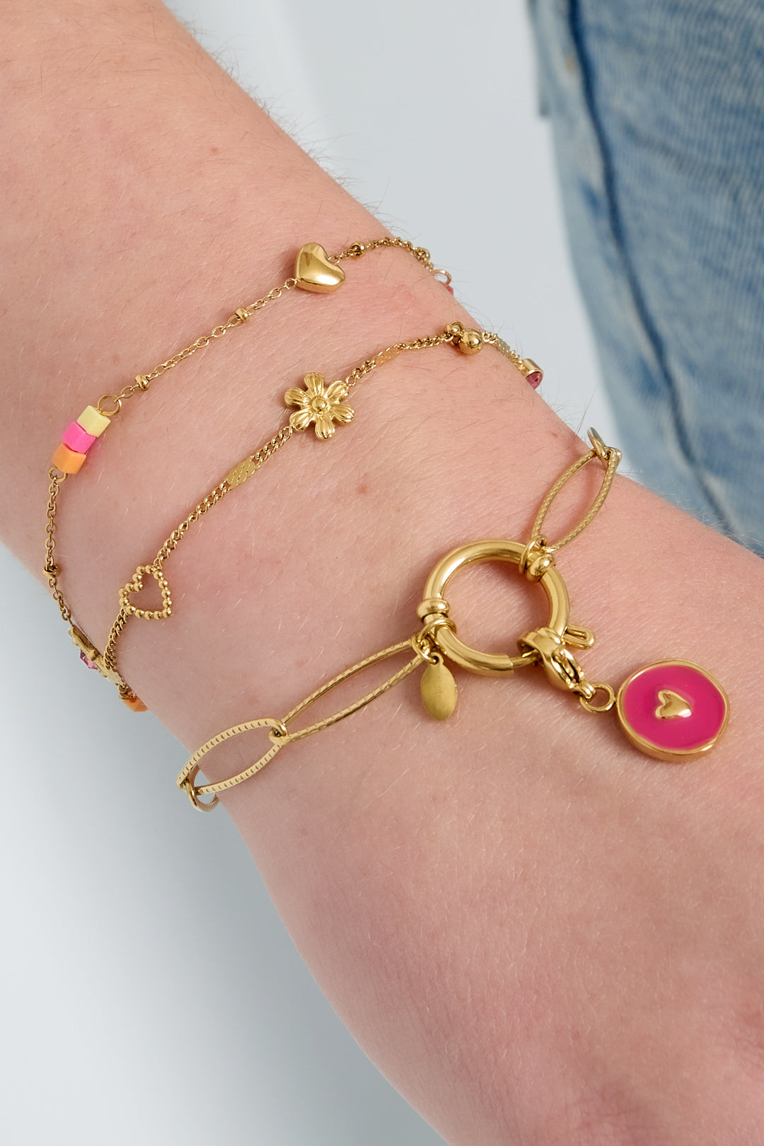 Close-up of a wrist wearing three gold bracelets including a playful charm bracelet with flower and heart details and colorful beads.