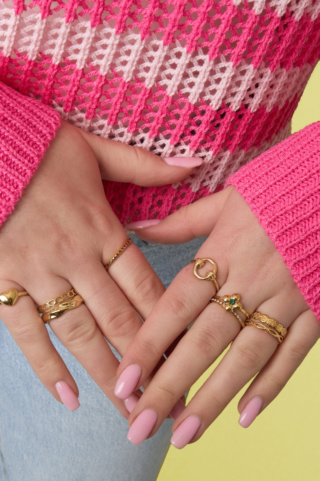 Woman's hands wearing multiple gold rings including a floral pattern gold ring, against a colorful pink and white knit sweater and light blue jeans