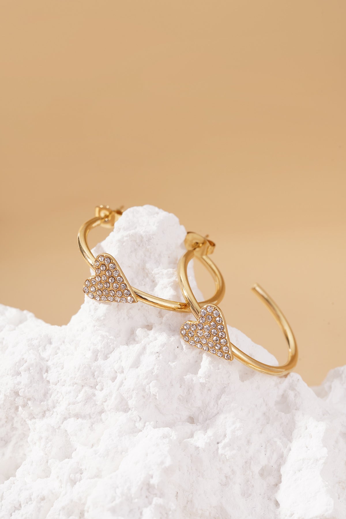 Gold hoop earrings featuring crystal-paved heart charms, styled on a white textured stone against a tan background.