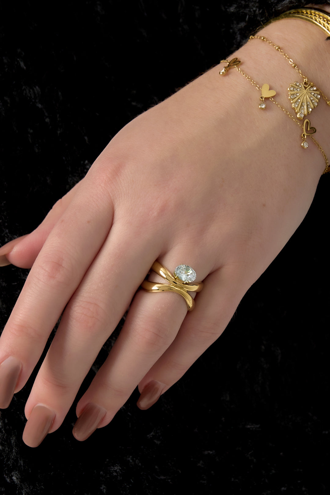 Female hand wearing the gold-tone twisted band ring with a sparkling oval stone paired with three delicate gold bracelets on a black background