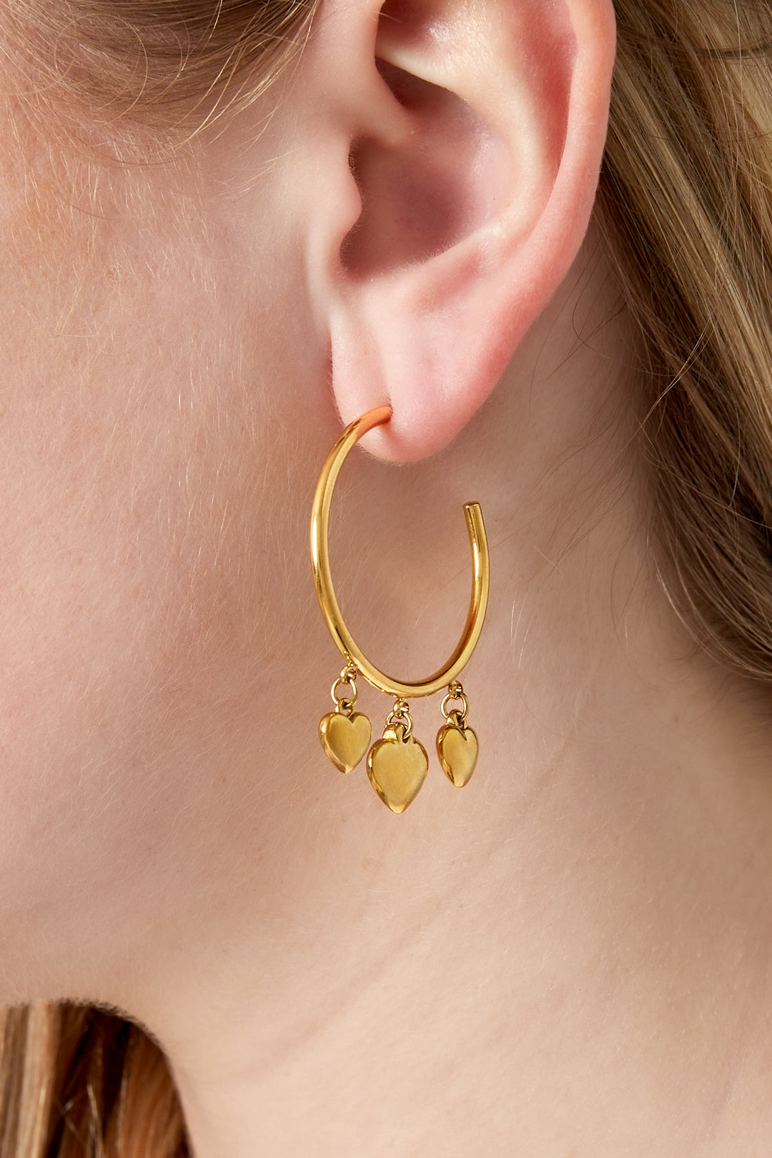 Close-up of a woman wearing a gold hoop earring with three dangling heart charms, showcasing the playful and minimal design.