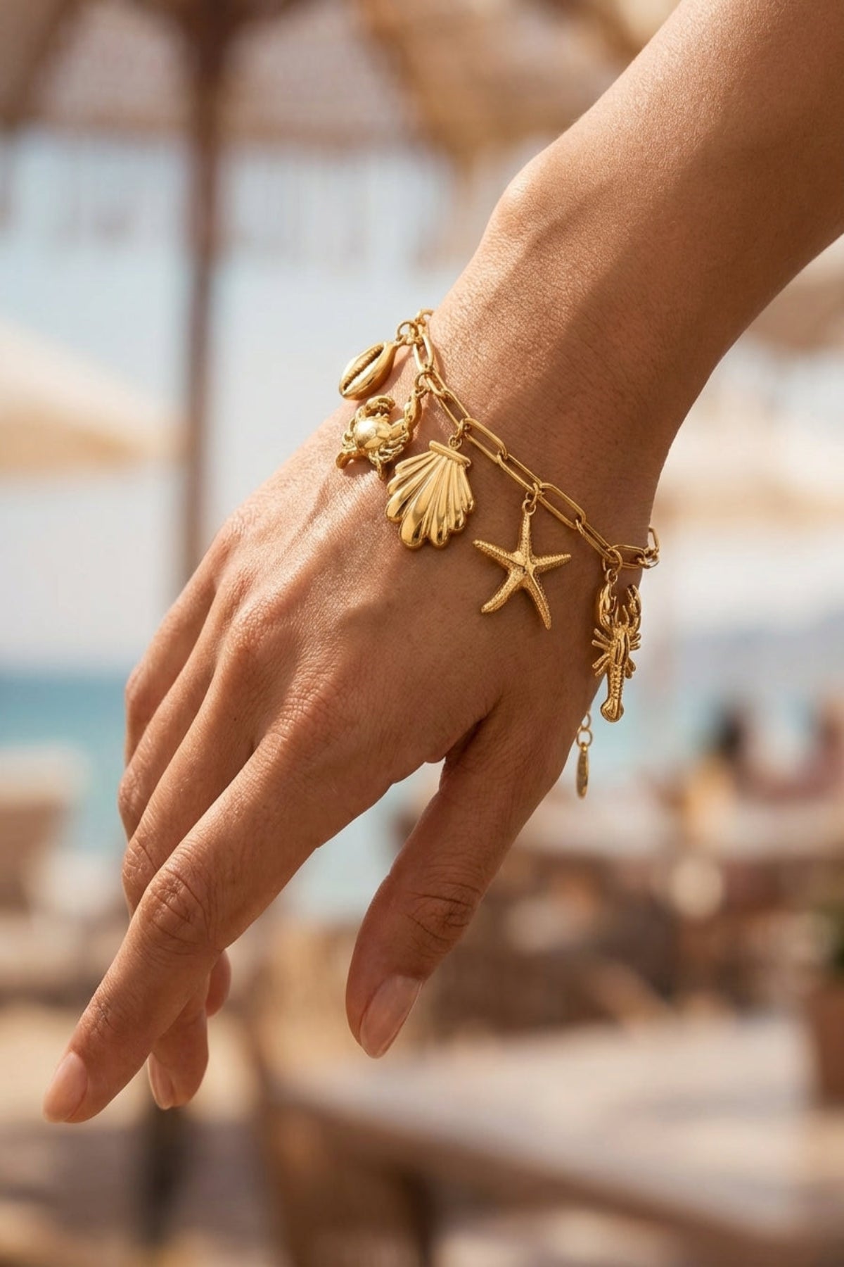 Hand wearing a gold bracelet with starfish and shell charms against a blurred beach background