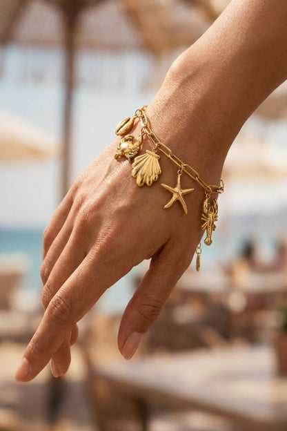 Hand wearing a gold bracelet with starfish and shell charms against a blurred beach background